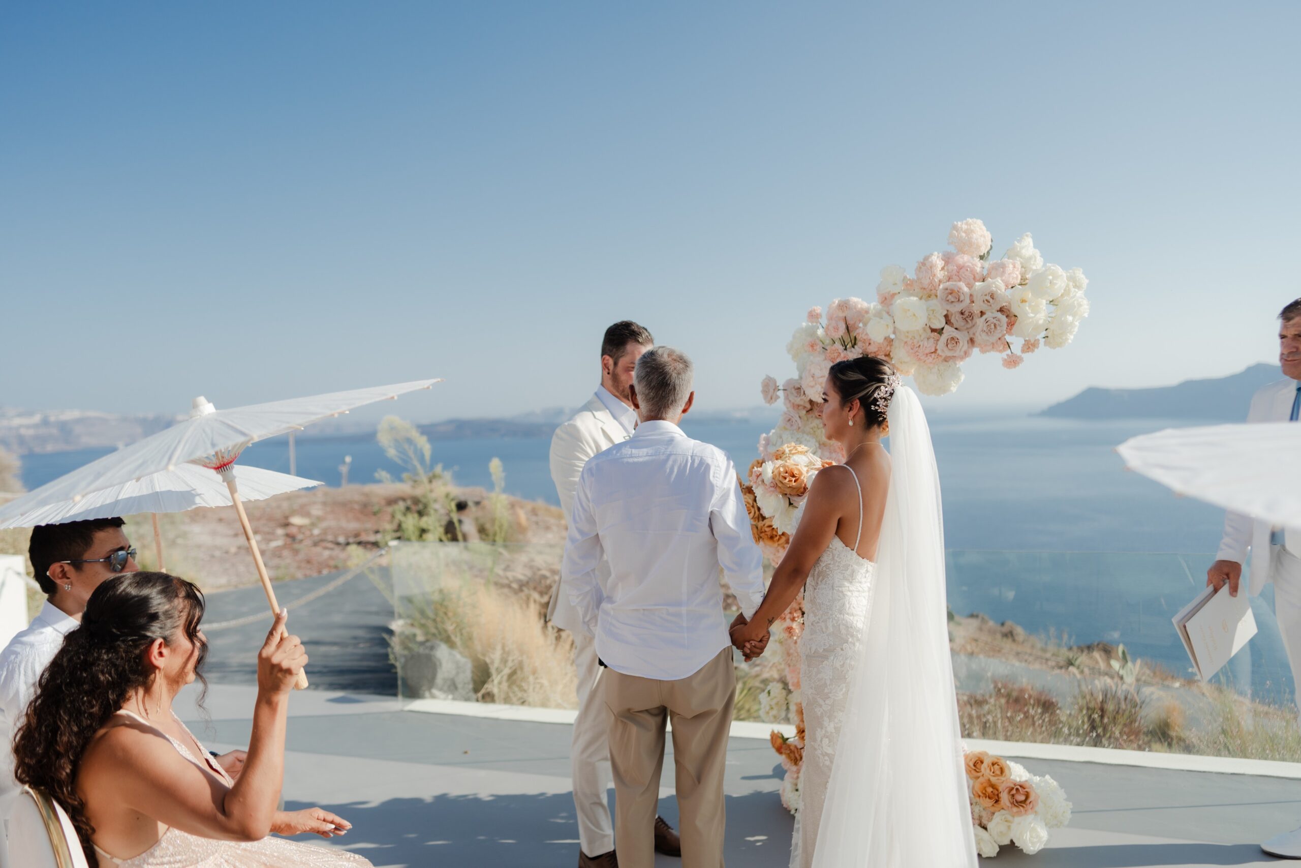 A couple stand before their celebrant at a clifftop ceremony, with parasols and a floral arch above the sea, Eventuries Wedding Planning and Styling