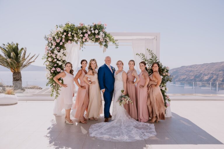 A bride, groom and bridesmaids in front of a floral arch on a terrace above the sea, Eventuries Wedding Planning and Styling