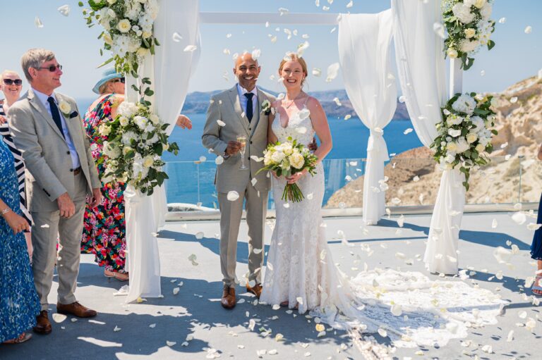 A couple walk down the aisle under white drapes and flowers as guests throw petals, with the sea behind, Eventuries Wedding Planning and Styling