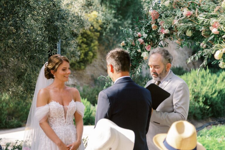 A bride and groom stand before the celebrant at an outdoor ceremony among olive trees and pink flowers, Eventuries Wedding Planning and Styling