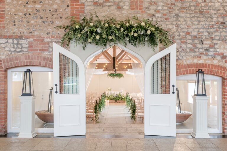Arched white doors in a flint wall open onto the Meadow Barn, set with chairs and greenery for a ceremony, Farbridge