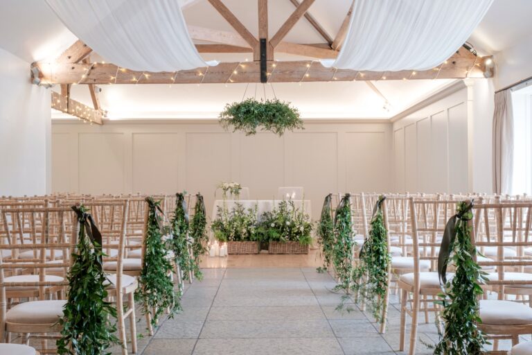 The aisle of the Meadow Barn, lined with foliage on the chairs, under draped fabric and a hanging floral hoop, Farbridge