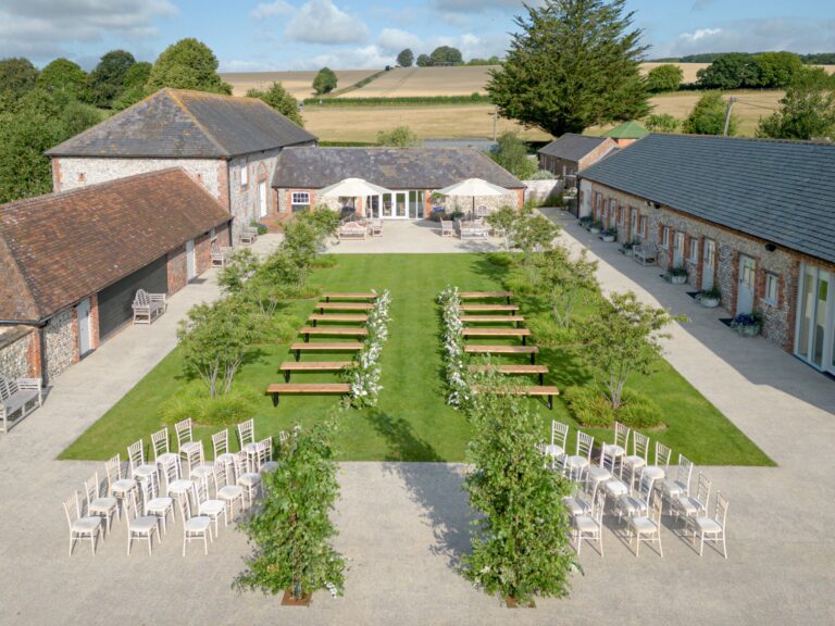 An outdoor ceremony on the courtyard lawn seen from above, with benches, chairs and flowering trees lining the aisle, Farbridge
