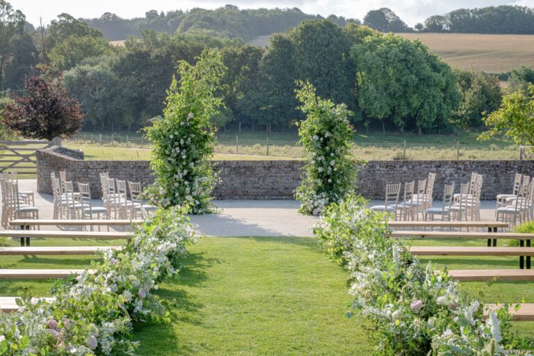 A flower-lined aisle across the courtyard lawn, with a flint wall and wooded downland beyond, Farbridge