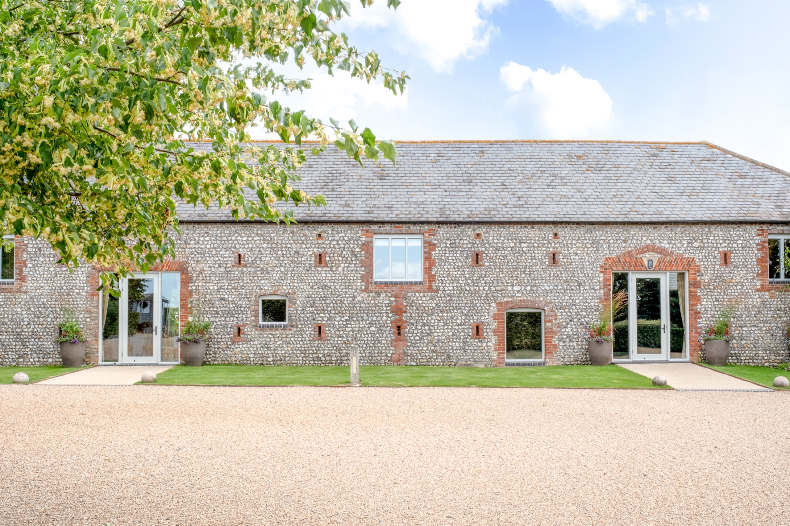 The long flint and brick front of the barn with glazed doors onto a gravel forecourt, Farbridge