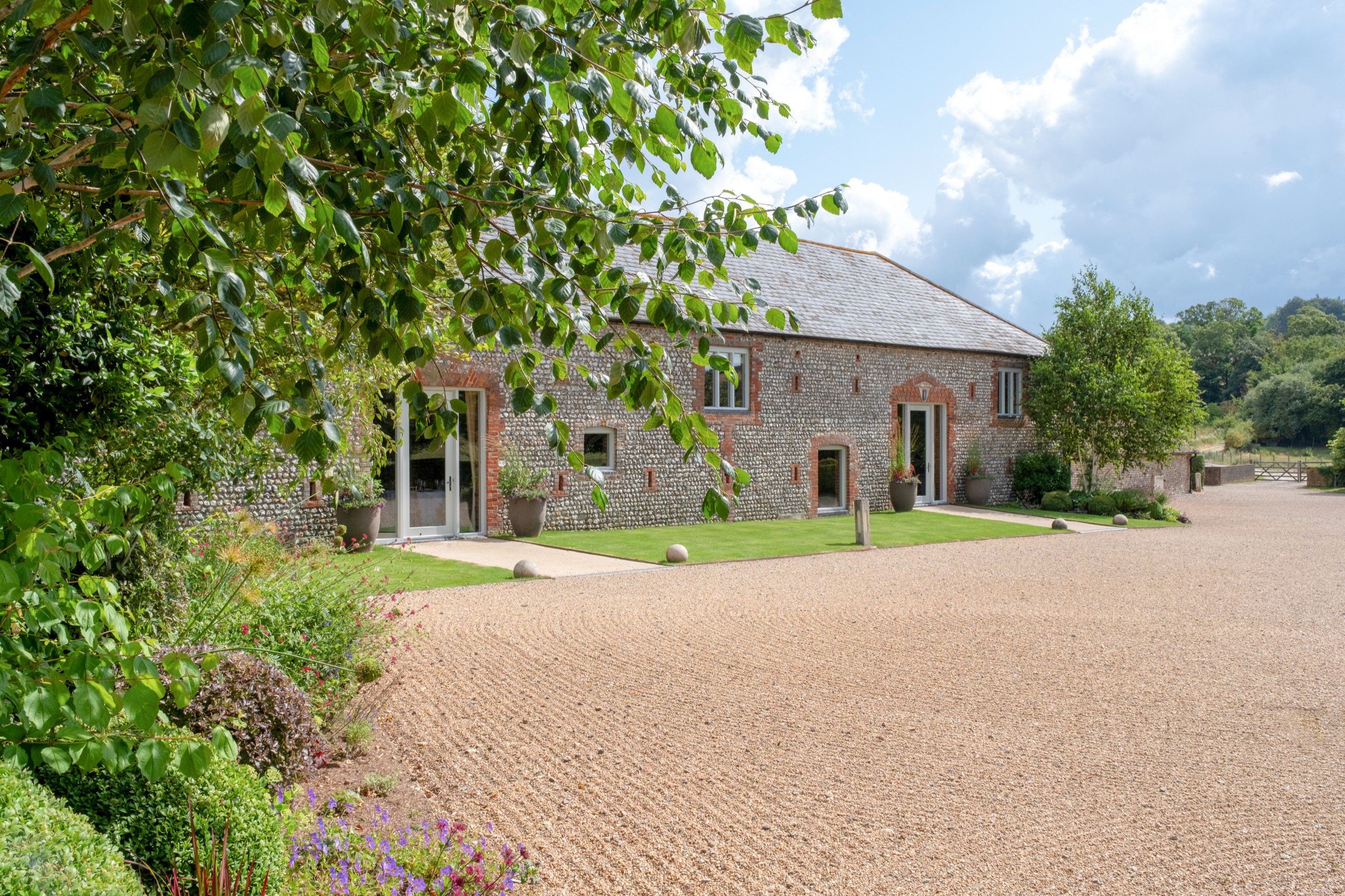 The barn range seen across the raked gravel drive, framed by trees and garden borders, Farbridge