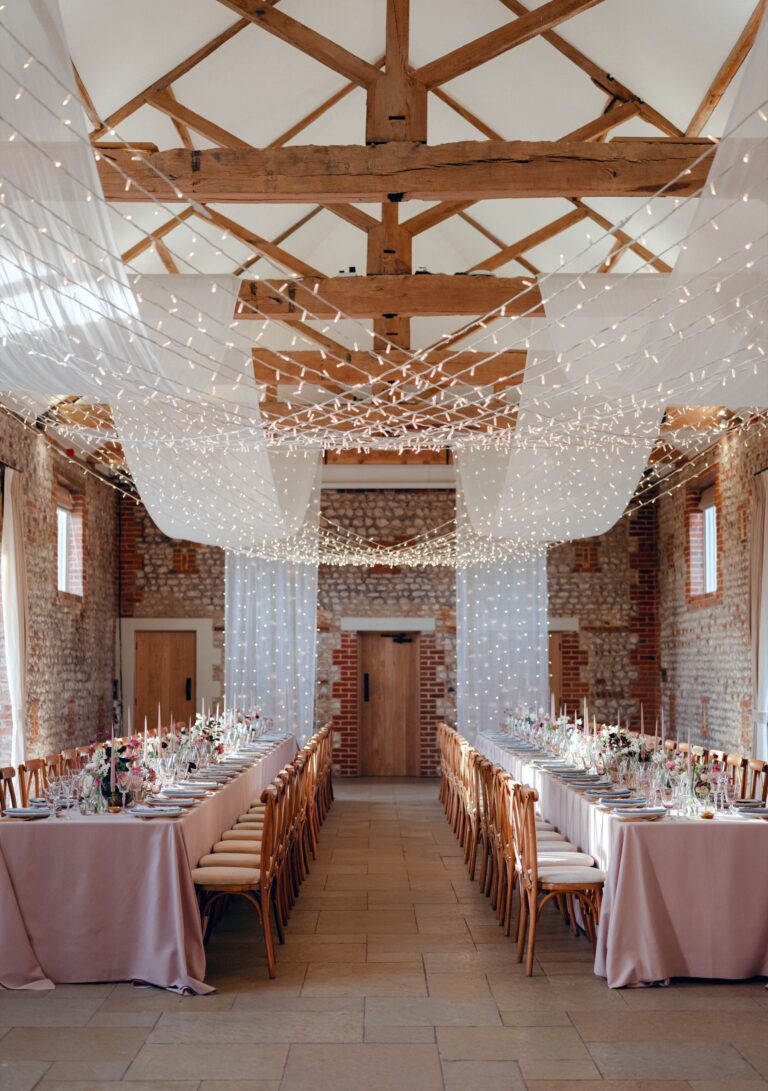 Long banqueting tables with blush linen beneath a canopy of fairy lights in the barn, Farbridge