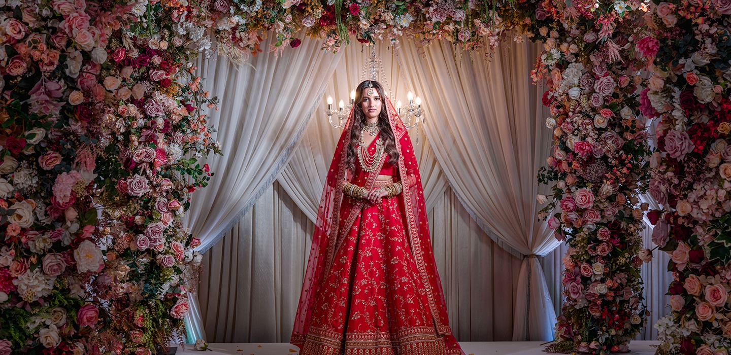 A bride in a red lehenga, beneath a floral arch, Farima Perry Florals and Events