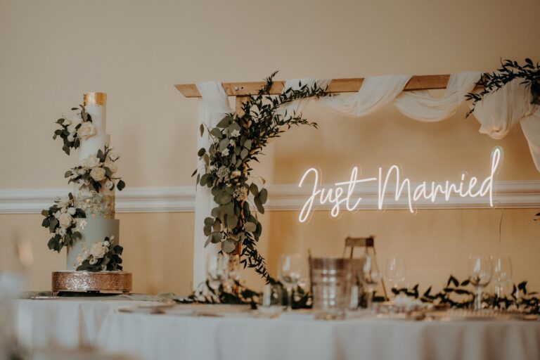 A four-tier cake beside a flower-wreathed frame with a Just Married neon sign, Farnham Castle