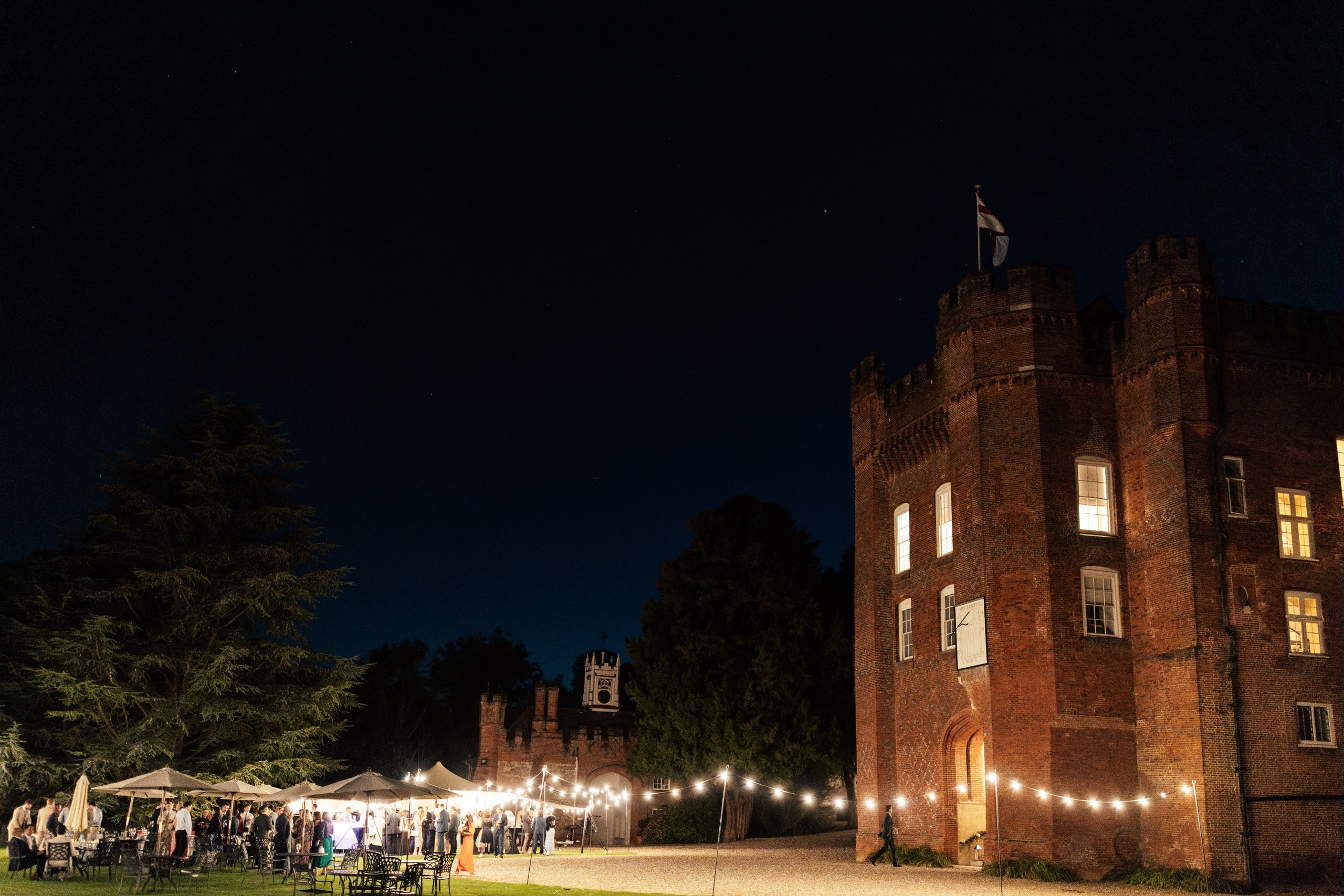 The entrance tower lit at night, with festoon lights strung across the gravel and a party under canopies on the lawn, Farnham Castle