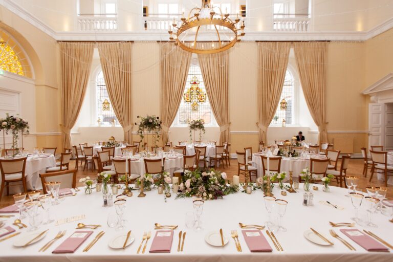 Round tables laid for a wedding breakfast in the Great Hall, with pink napkins, gold cutlery and a stained-glass window behind, Farnham Castle