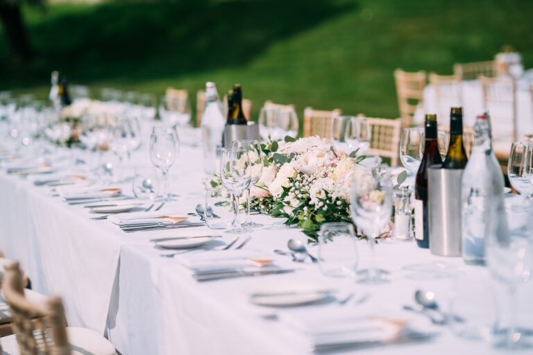 A long table on the East Lawn laid for an outdoor meal, with white flowers, wine and glassware, Farnham Castle