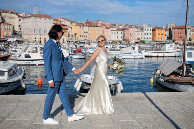 The couple walk hand in hand along the harbour front with colourful houses behind, Rovinj, Flammeum