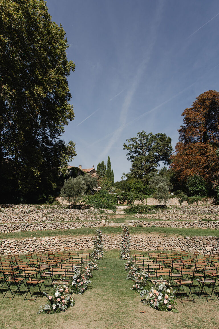 Wooden chairs set out for a ceremony in a terraced garden with floral arrangements along the aisle, Fleurs design By Faustine