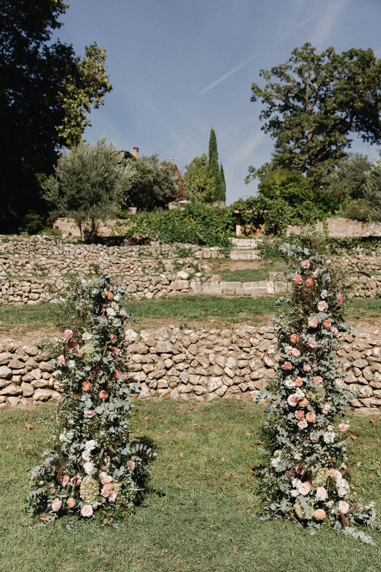 Two tall floral columns framing a ceremony space on a stone-walled garden terrace, Fleurs design By Faustine