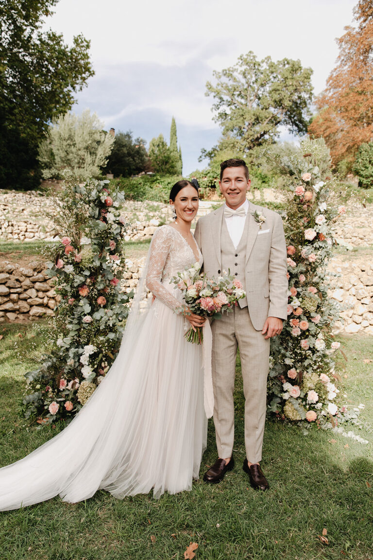 A couple standing between two tall floral columns at their garden ceremony, Fleurs design By Faustine