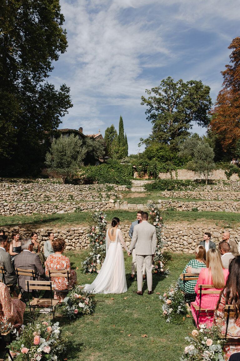 The aisle at a garden ceremony with the couple facing the guests and floral arrangements along the path, Fleurs design By Faustine