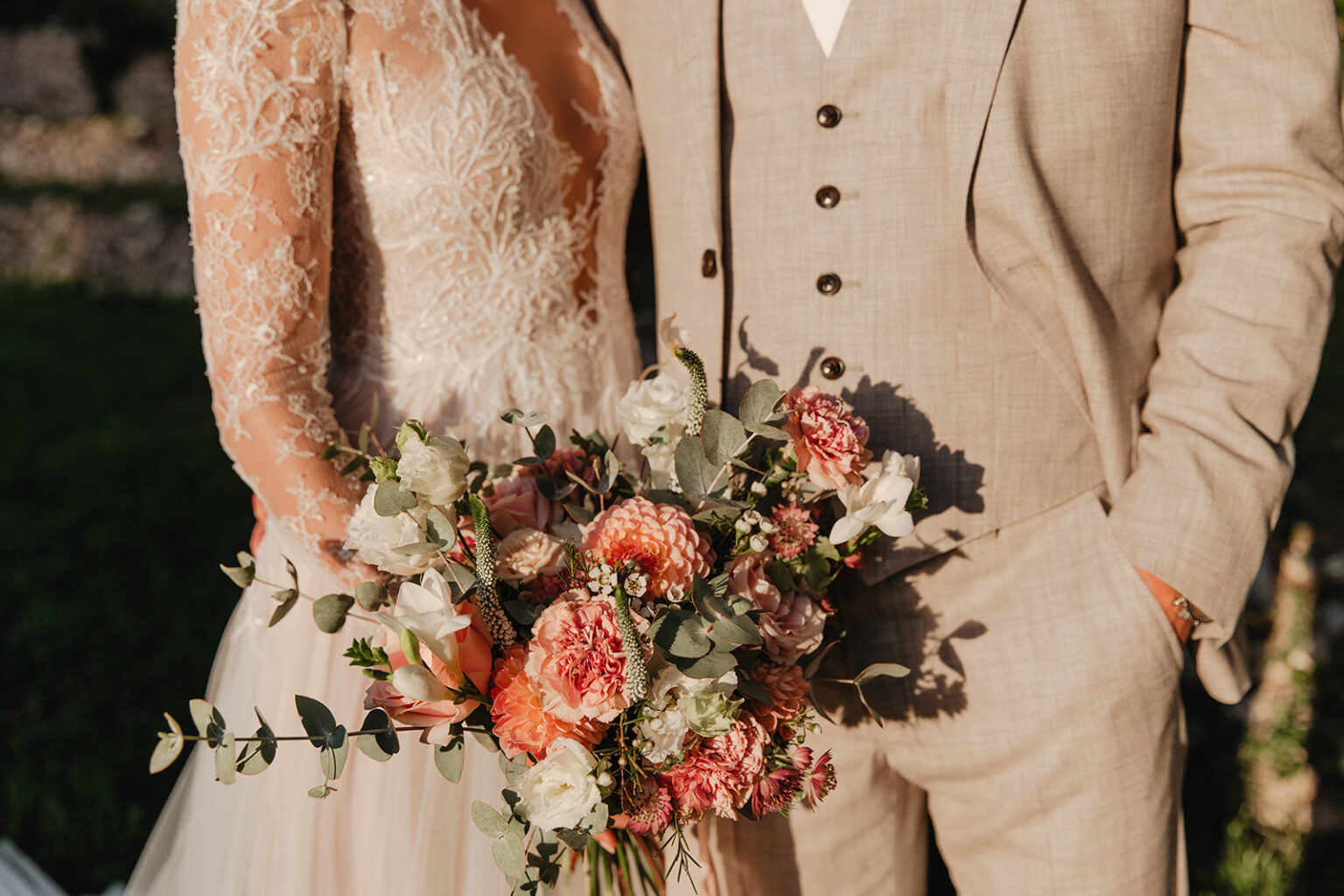 A bride and groom in neutral tones with a loose bouquet of peach, coral and cream roses