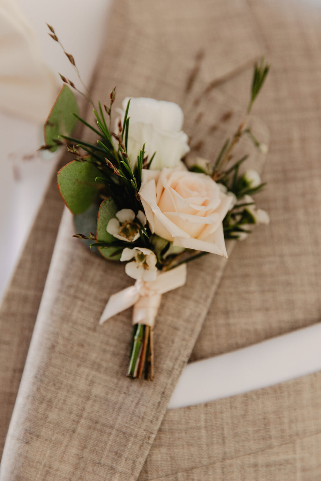 A boutonnière of a cream rose with foliage and a ribbon on a linen jacket lapel