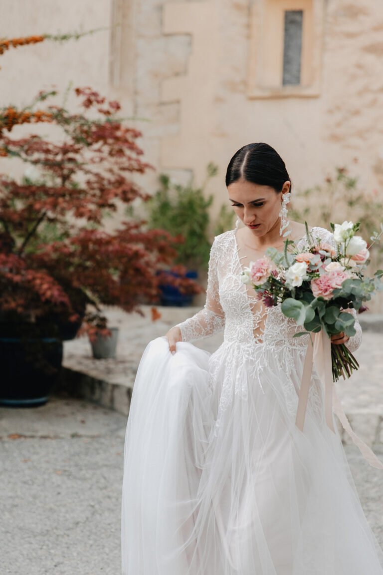 A bride in a lace gown walking across a courtyard holding a soft pink and white bouquet, Fleurs design By Faustine