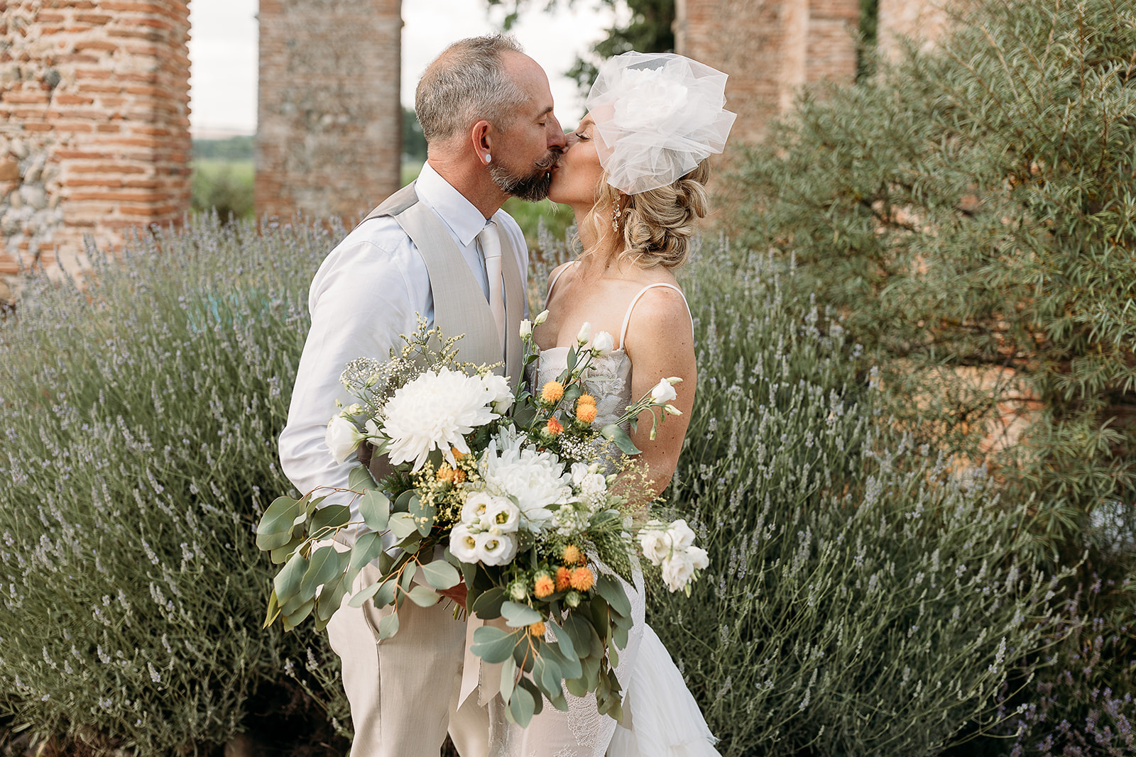 A couple kissing among lavender, the bride holding a white bouquet with orange and greenery, Fleurs design By Faustine