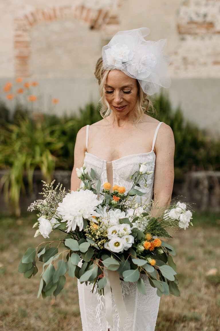 A bride with a feathered headpiece holding a white and orange bouquet in front of old brick walls, Fleurs design By Faustine
