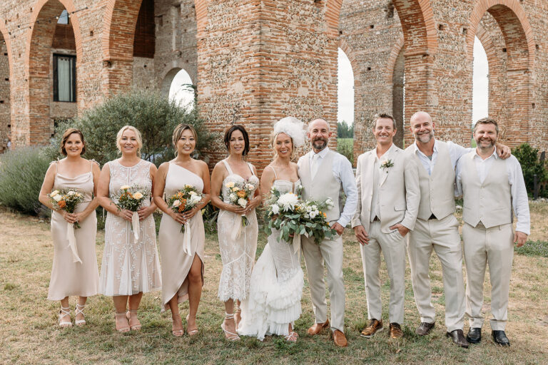 A wedding party in neutral tones holding bouquets in front of brick arches, Fleurs design By Faustine