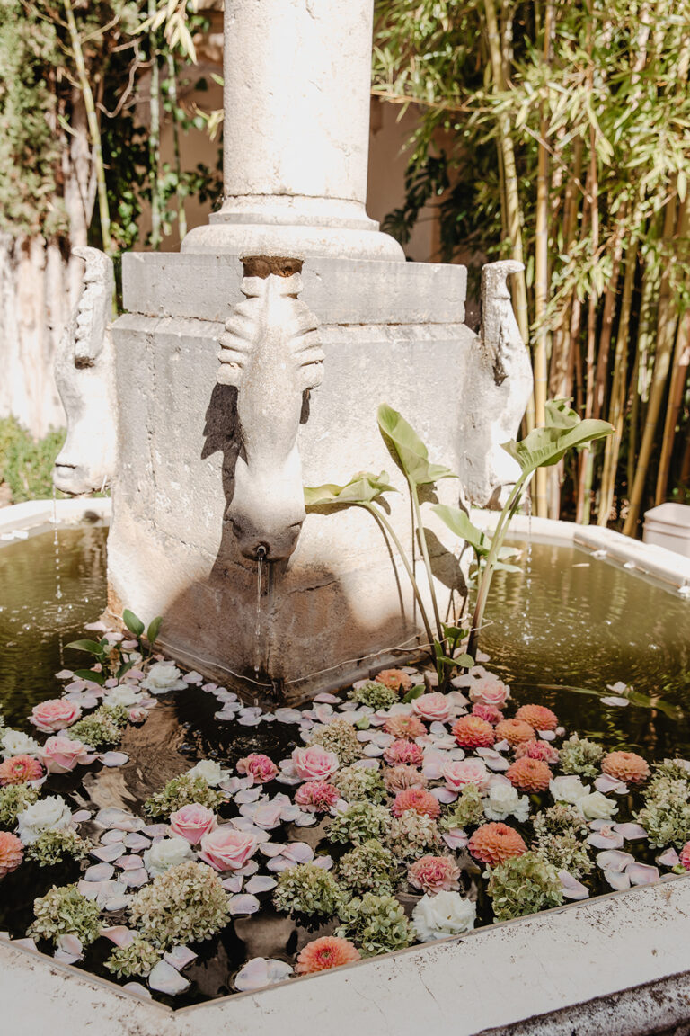 A stone fountain in a garden with a basin of floating pink, peach and green blooms, Fleurs design By Faustine