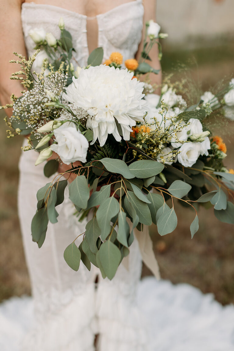 A bridal bouquet of white dahlias, orange accents and eucalyptus held against a white gown, Fleurs design By Faustine
