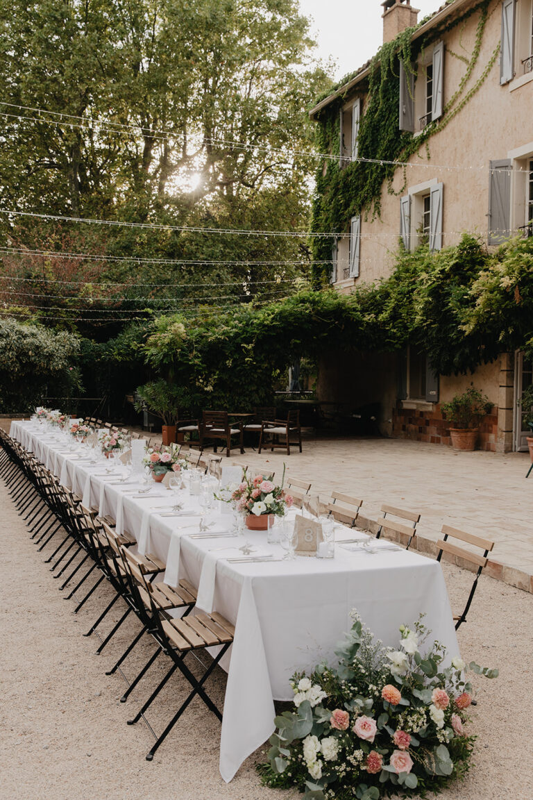 A long reception table dressed in white linen in a courtyard beside a stone house, with a floor arrangement at the end, Fleurs design By Faustine