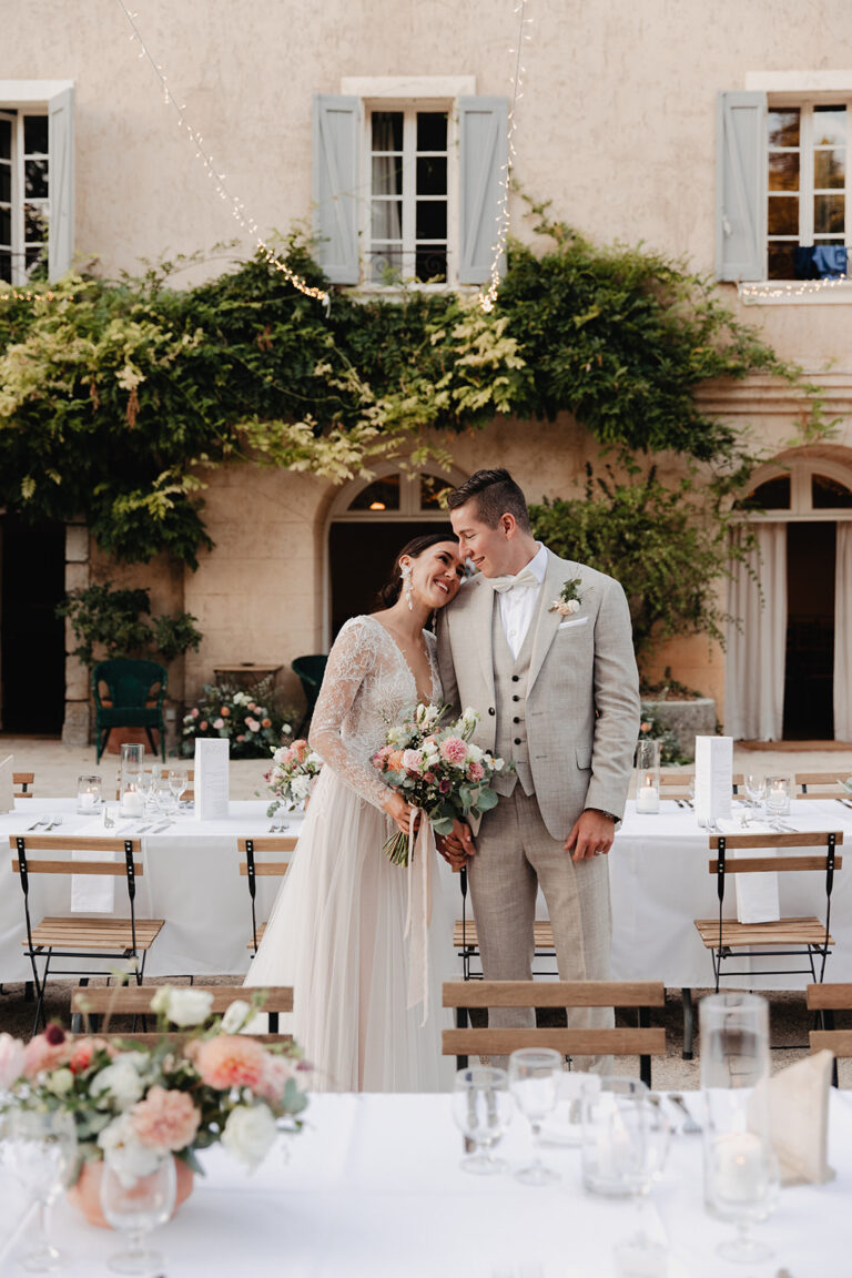 A couple embracing at a courtyard dinner table with a low floral centrepiece in the foreground, Fleurs design By Faustine