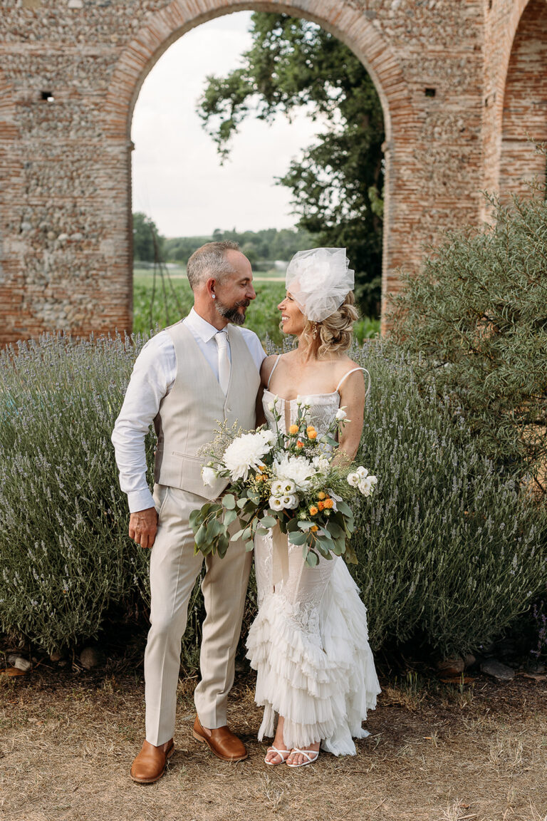 A couple standing before a brick arch in old ruins, the bride holding a white bouquet, Fleurs design By Faustine