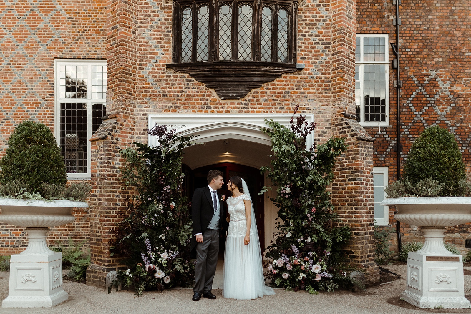A couple embracing beneath the Tudor Courtyard entrance archway, red brick framing them and florals to either side, Fulham Palace