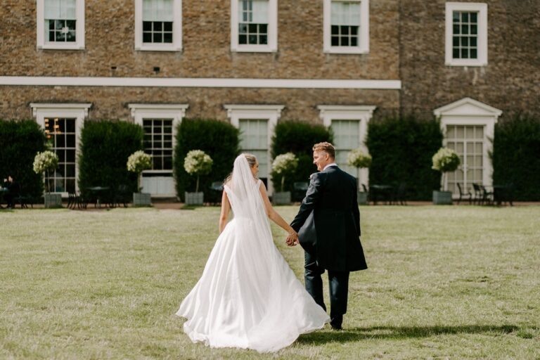 A couple walking hand in hand across the back lawn, the east front of the palace behind them, Fulham Palace