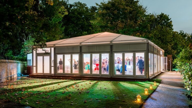 The Chaplain's Marquee lit from within at dusk, its glass walls glowing on the lawn, Fulham Palace