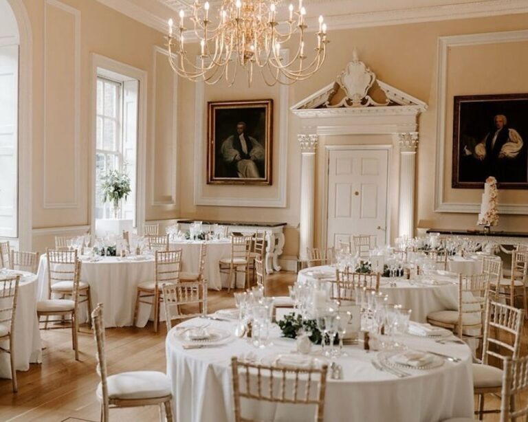 A palace state room set for a wedding breakfast, round tables and a chandelier beneath hung portraits, Fulham Palace
