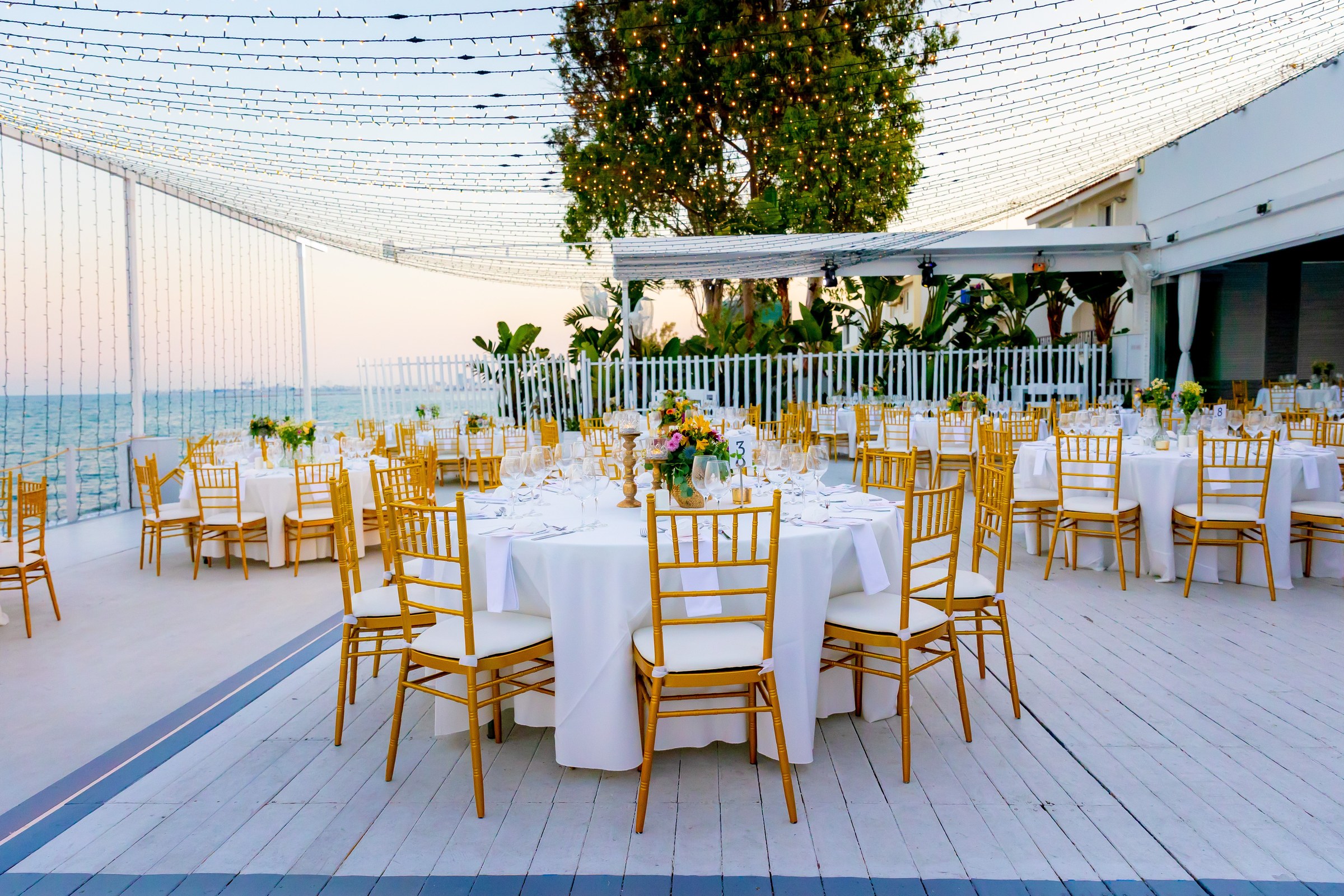 Outdoor deck set for a wedding dinner beneath a canopy of lights beside the white seafront building, Galu Seaside