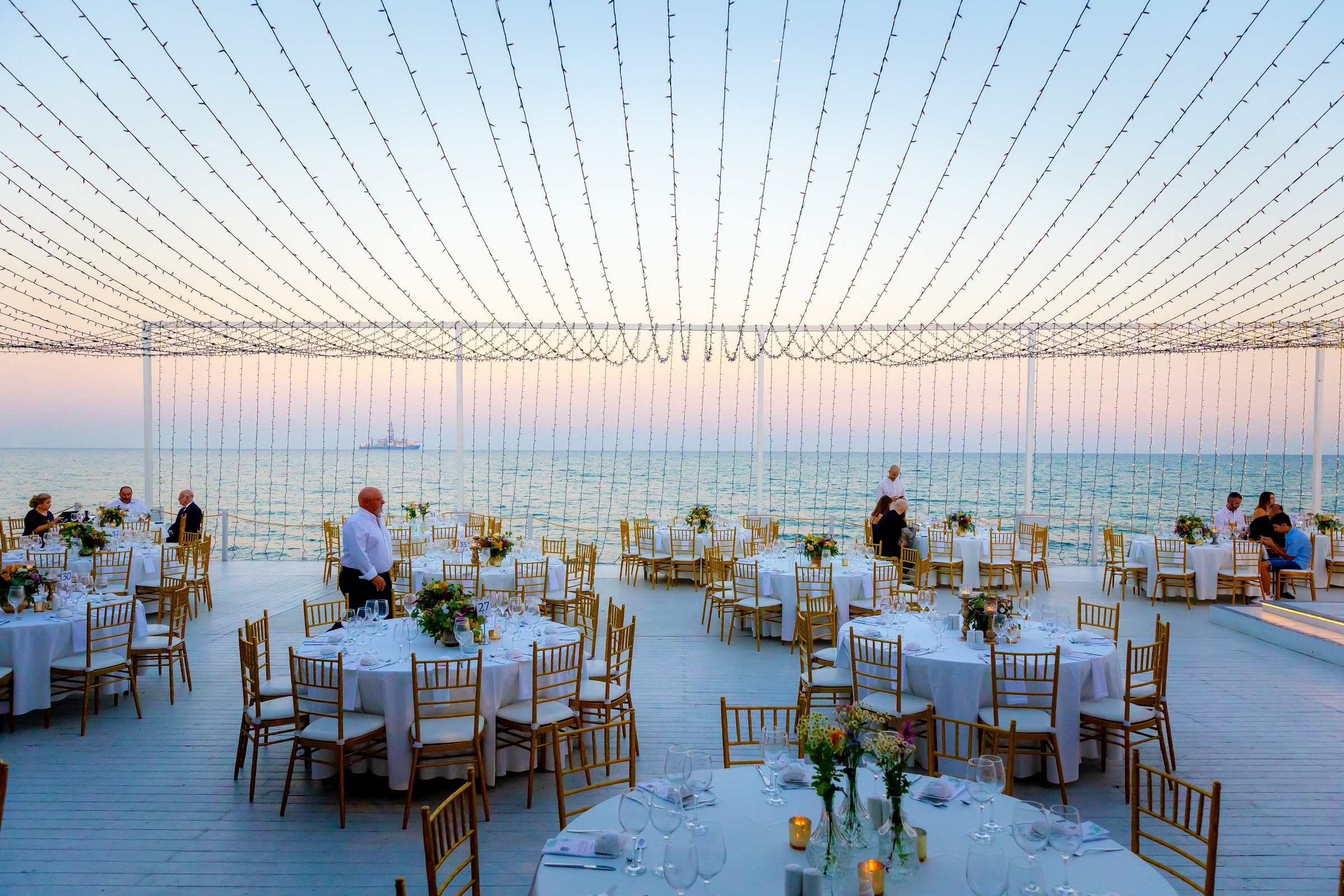 Round tables on the deck at sunset under the canopy of lights, with the sea behind, Galu Seaside