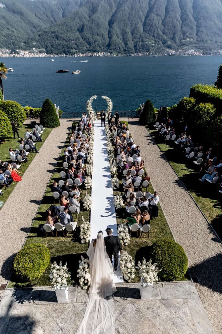 Aerial view of a garden ceremony on a lakeside terrace with the bride walking the aisle, Geta Ungurean Agency