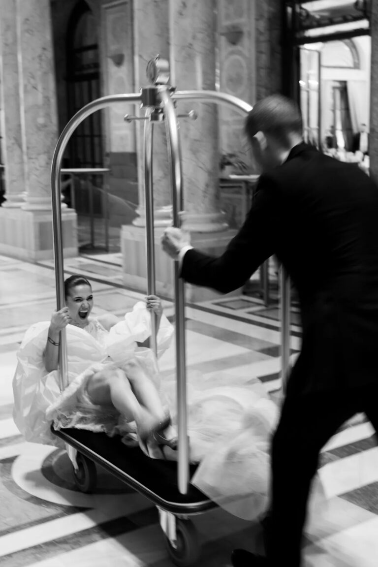 Black and white image of a bride riding a hotel luggage trolley, Geta Ungurean Agency