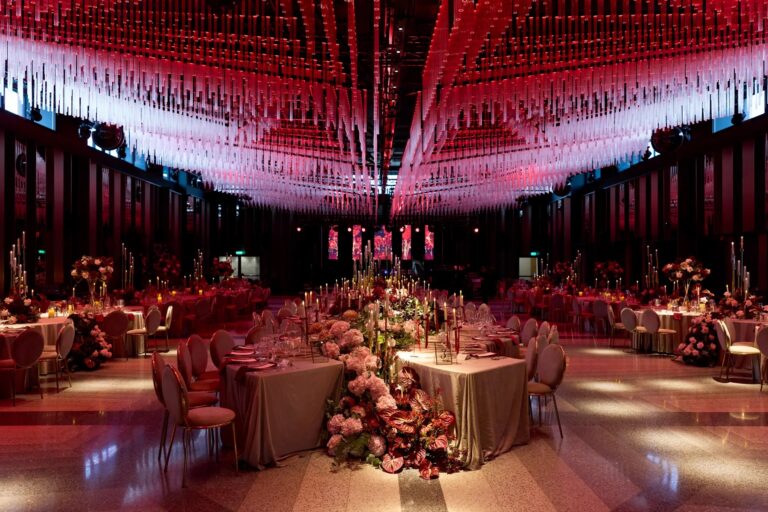 Reception hall with a suspended red and pink ceiling installation and long floral tables, Geta Ungurean Agency