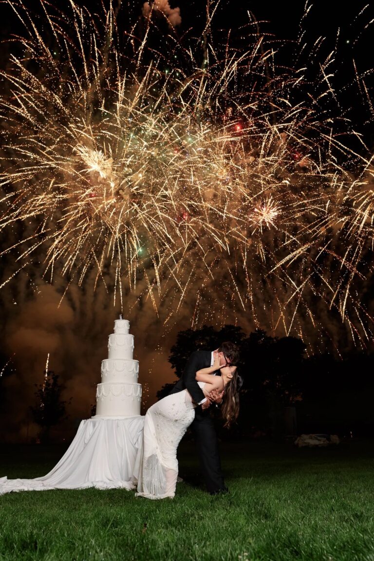 Couple kissing beside a tiered white cake as fireworks light the sky, Geta Ungurean Agency