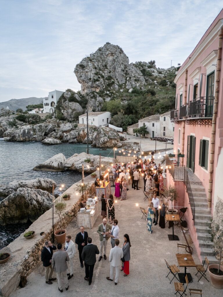 Guests gathering at dusk on a waterfront lane strung with lights in a seaside village, a great rock rising behind, Giandomenico Cosentino Photography