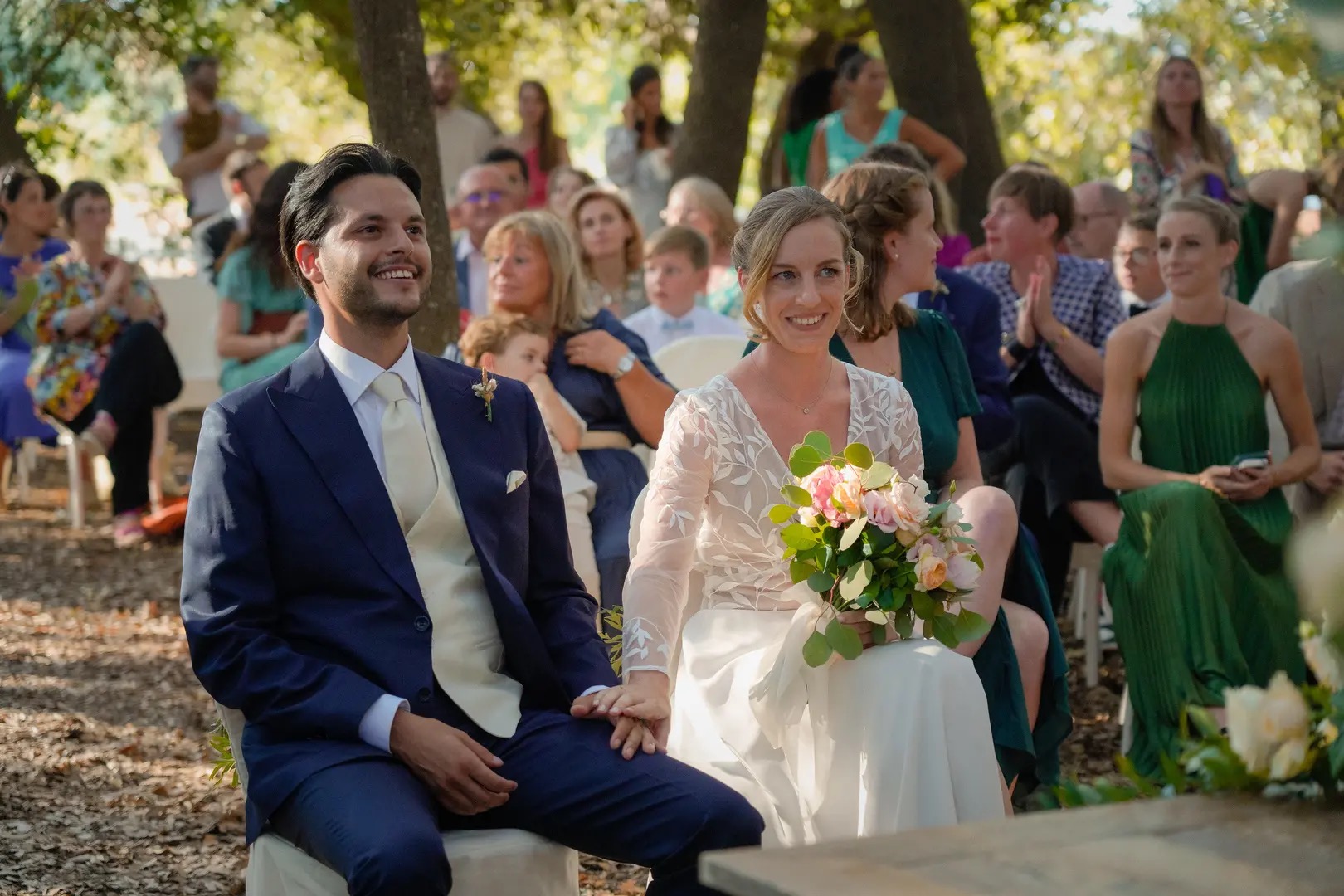 The couple seated at their outdoor ceremony with guests close behind, Puglia, Giuseppe Suma Weddings