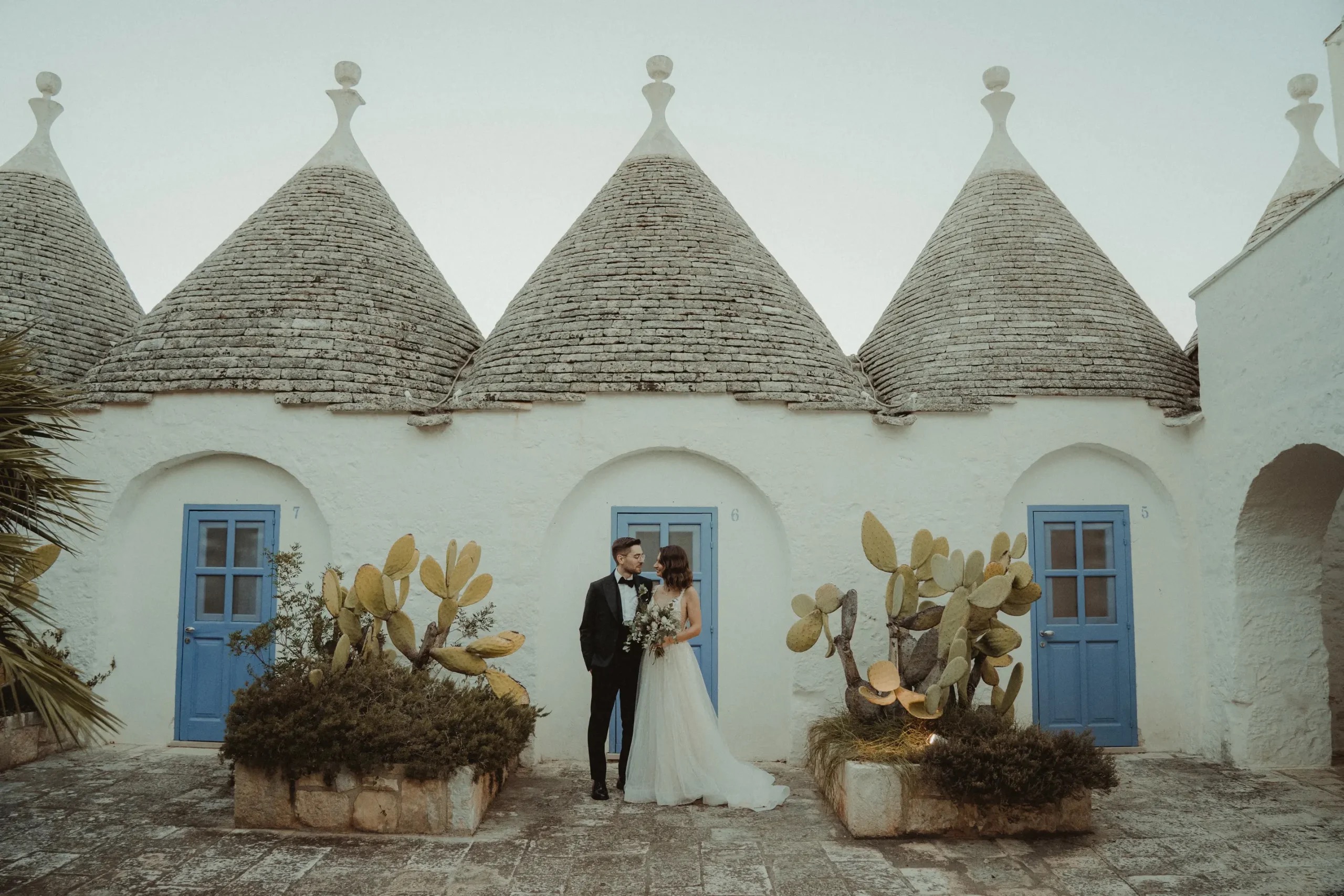 A couple outside a whitewashed trullo with blue doors and prickly pears, Valle d'Itria, Puglia, Giuseppe Suma Weddings