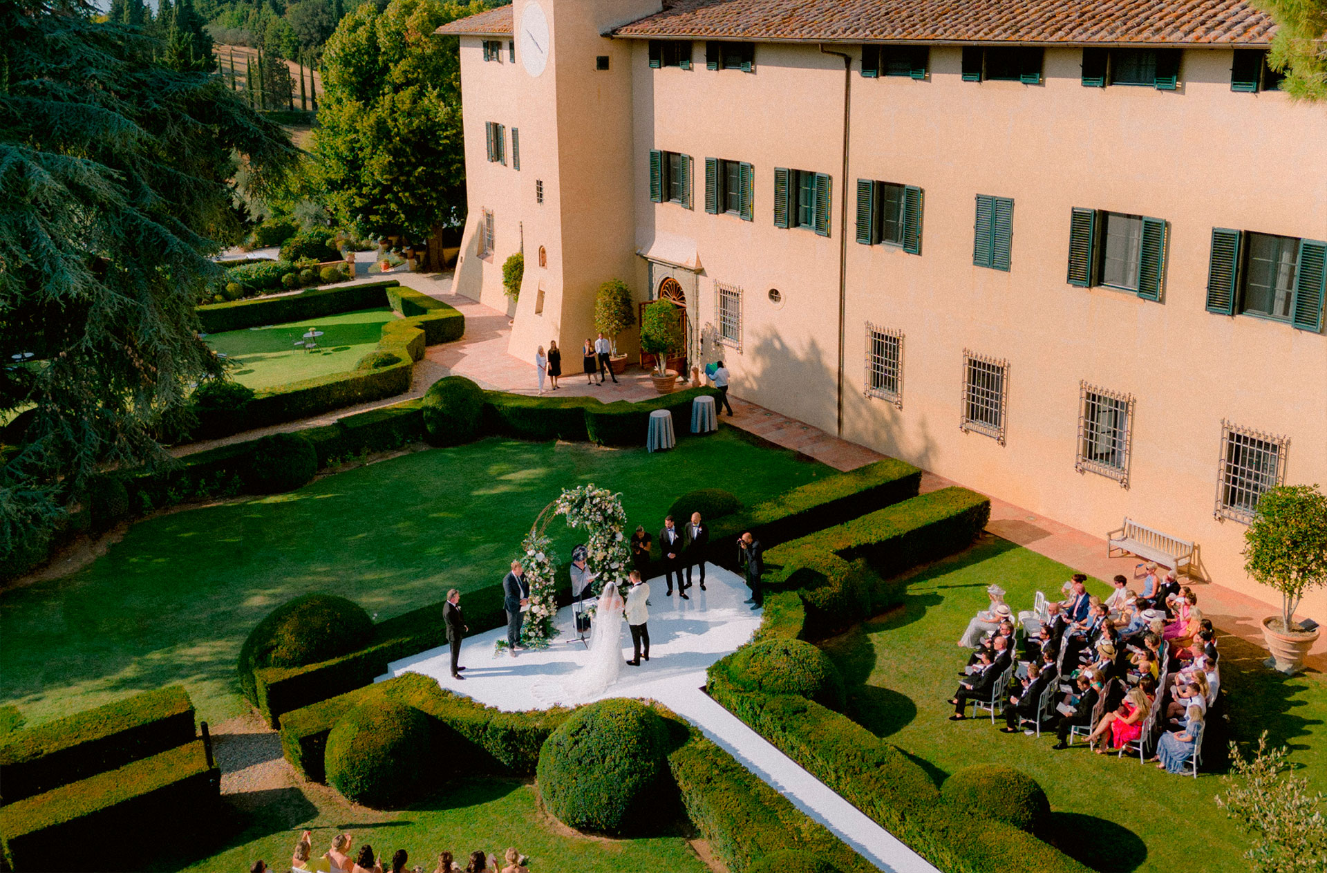 From above: a ceremony on a white circular platform in a villa's box-hedge garden in Tuscany, guests seated on the lawn, Glam Events in Tuscany