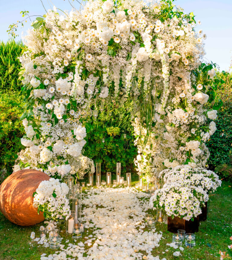 An arch of white flowers and hanging greenery over a petal-strewn path in a garden in Tuscany, Glam Events in Tuscany