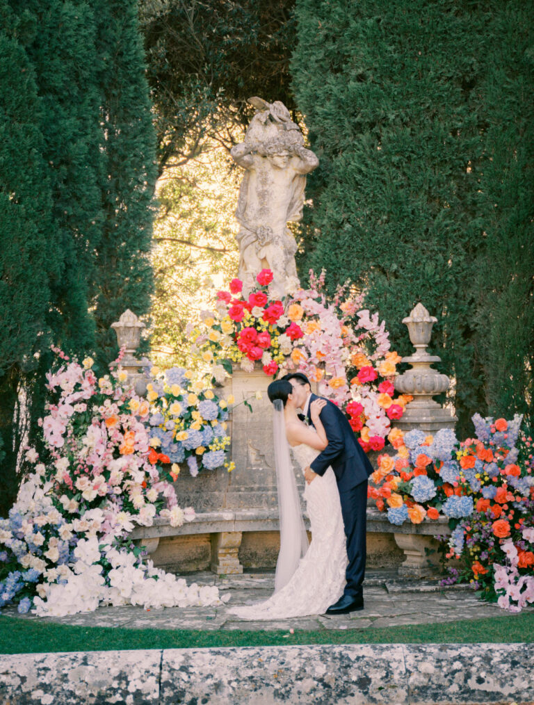 A couple at a garden fountain dressed with bright roses, orchids and hydrangeas, cypress hedges either side, Glam Events in Tuscany
