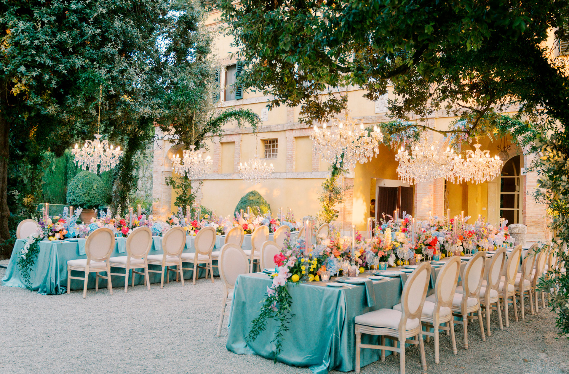 Dinner in a villa courtyard in Tuscany: crystal chandeliers hung from the trees over long tables in teal linen with bright mixed flowers, Glam Events in Tuscany