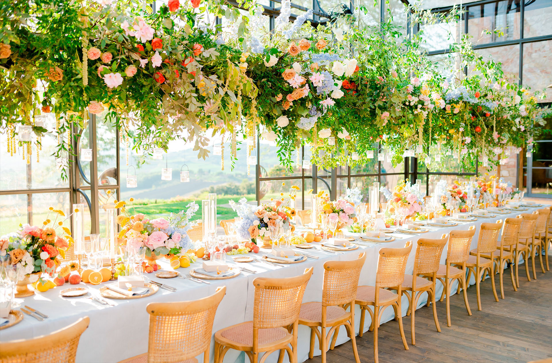 A long table in a glass pavilion under a hanging installation of roses and greenery, the Tuscan hills through the windows, Glam Events in Tuscany